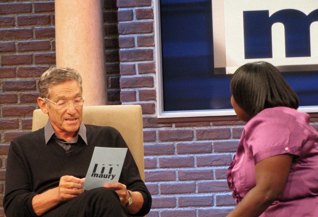 TV personality Maury Povich on the set of his talk show, sitting in a chair and engaging with a woman.