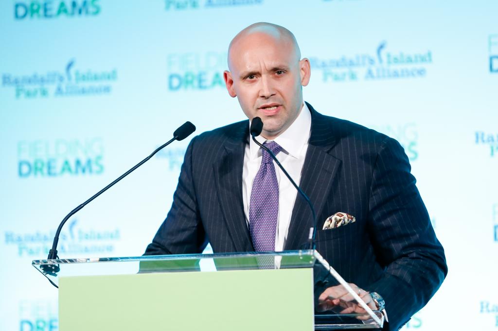 Justin Hamill in a suit speaking in front of a podium.