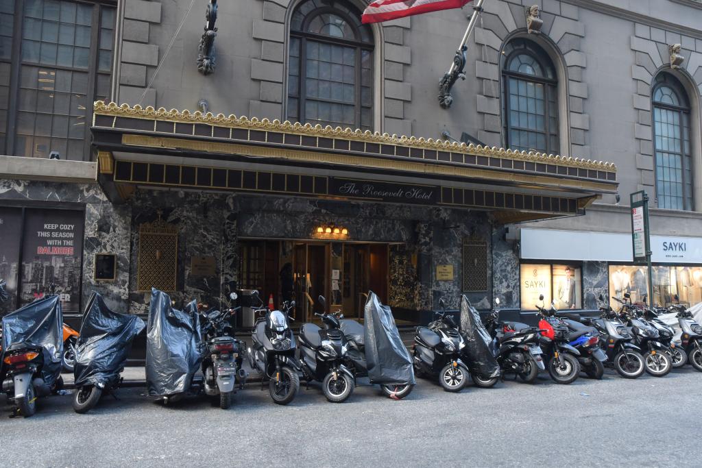 A row of motorized scooters parked on a city block in front of the Roosevelt Hotel on Madison Ave and E 46th Street.