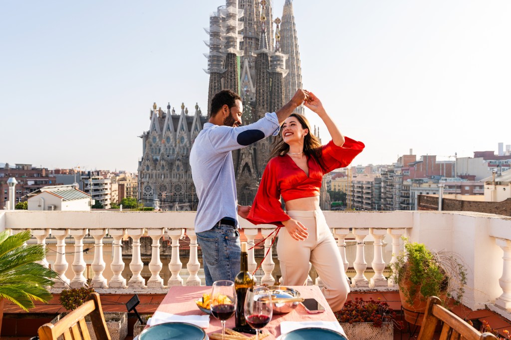 Multiracial couple happily enjoying a romantic dinner on a rooftop overlooking Sagrada Familia, Barcelona