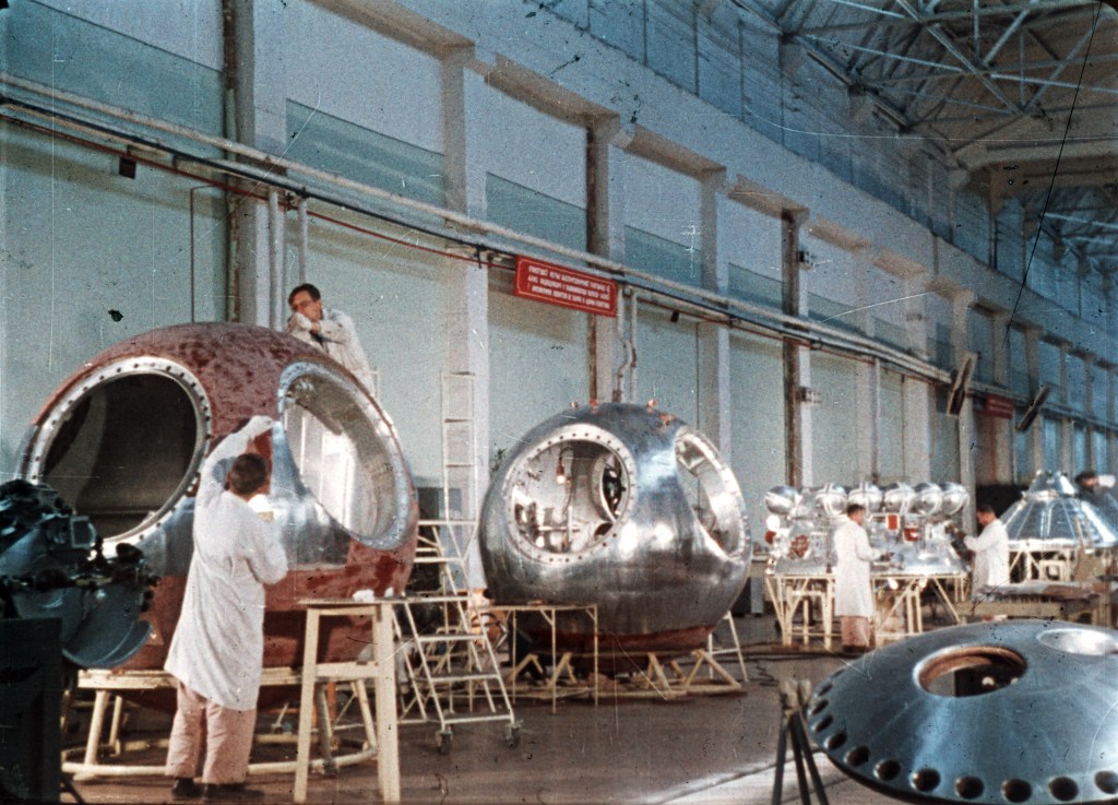 A Russian scientist (face unseen) works on the Vostok 1, with others scientists and space gadgets in the backgroun. 