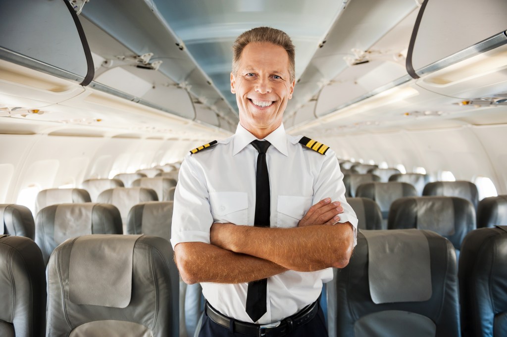 Confident male pilot in uniform, arms crossed, smiling inside of an airplane