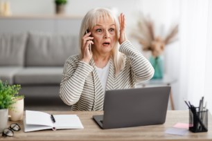 Surprised mature woman talking on mobile phone with a look of shock, seated at desk with laptop