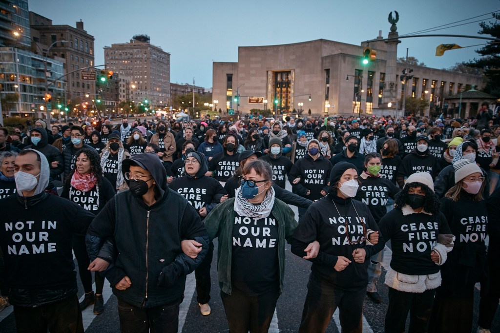 Protesters with shirts reading "Cease fire now" and "Not in our name" fill up a Brooklyn street, blocking traffic. 