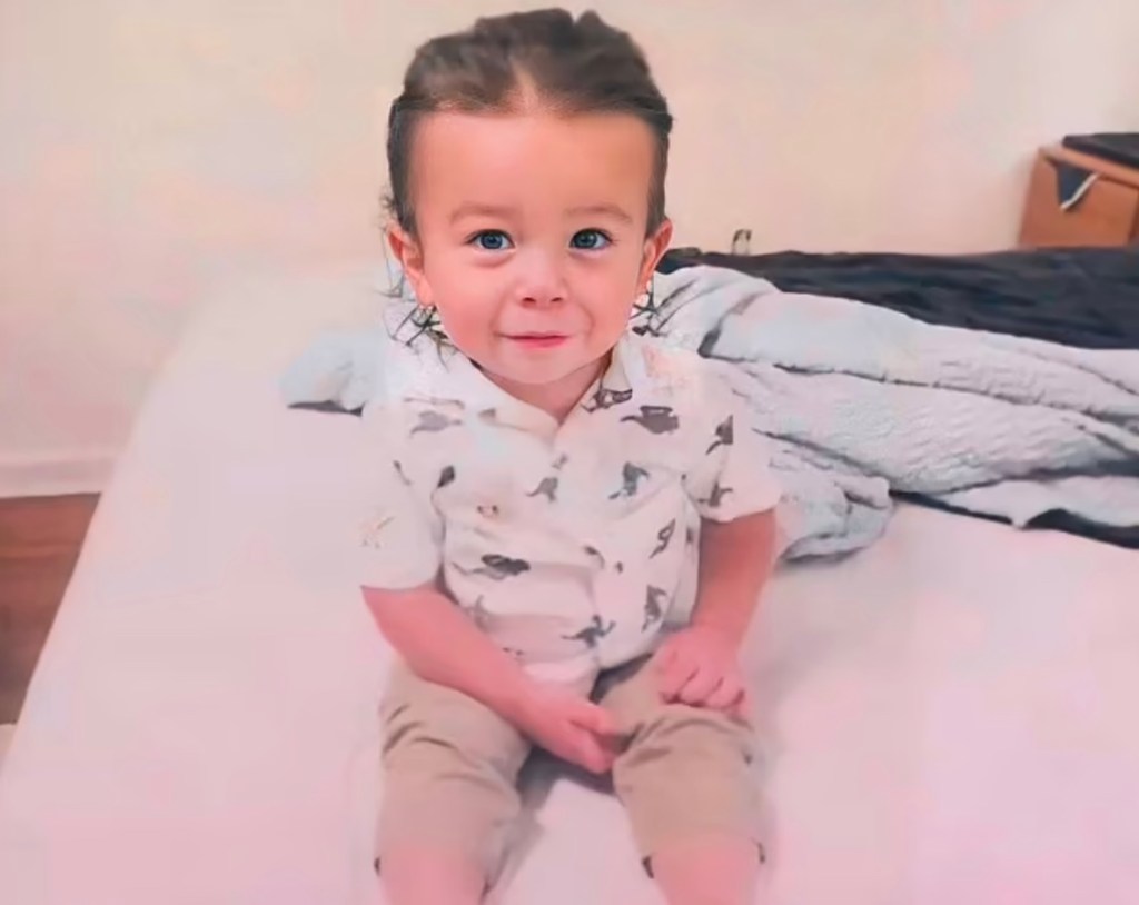 Quinton Simon wears a collared shirt and shorts and smiles while sitting on a bed with his hair combed back