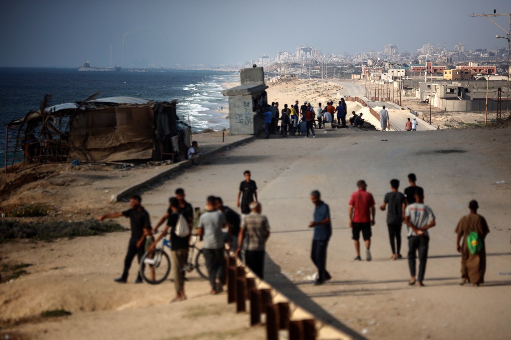 More than a dozen residents of Gaza gather near the shore, where a ship can be seen in the distance.