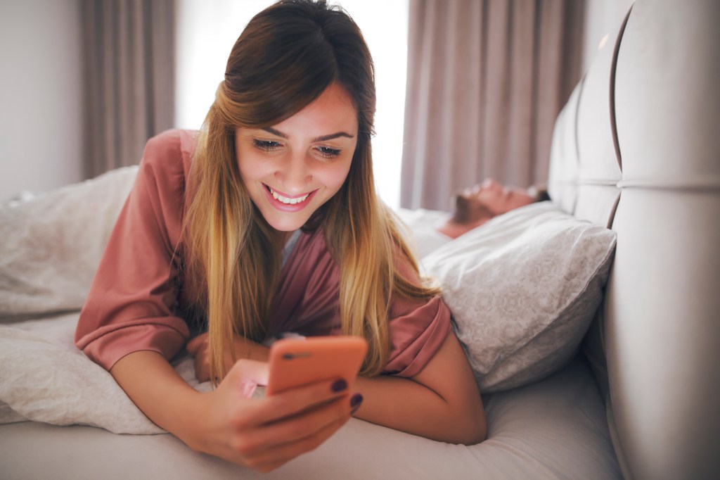 Smiling woman reading text message while her partner are sleeping