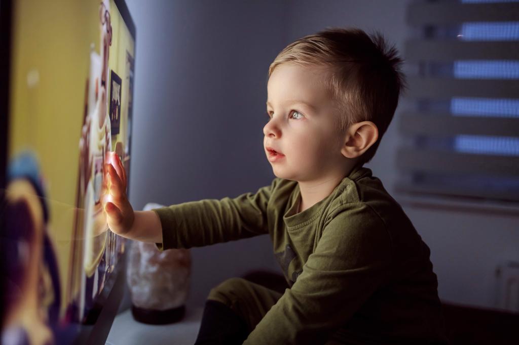 Young boy in pajamas sitting close to the TV and touching the screen while watching a cartoon