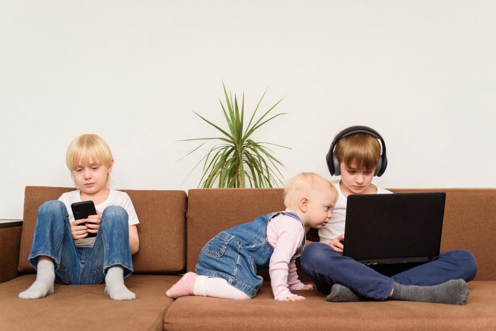 Three children sitting on a sofa, engrossed in using a laptop and a smartphone