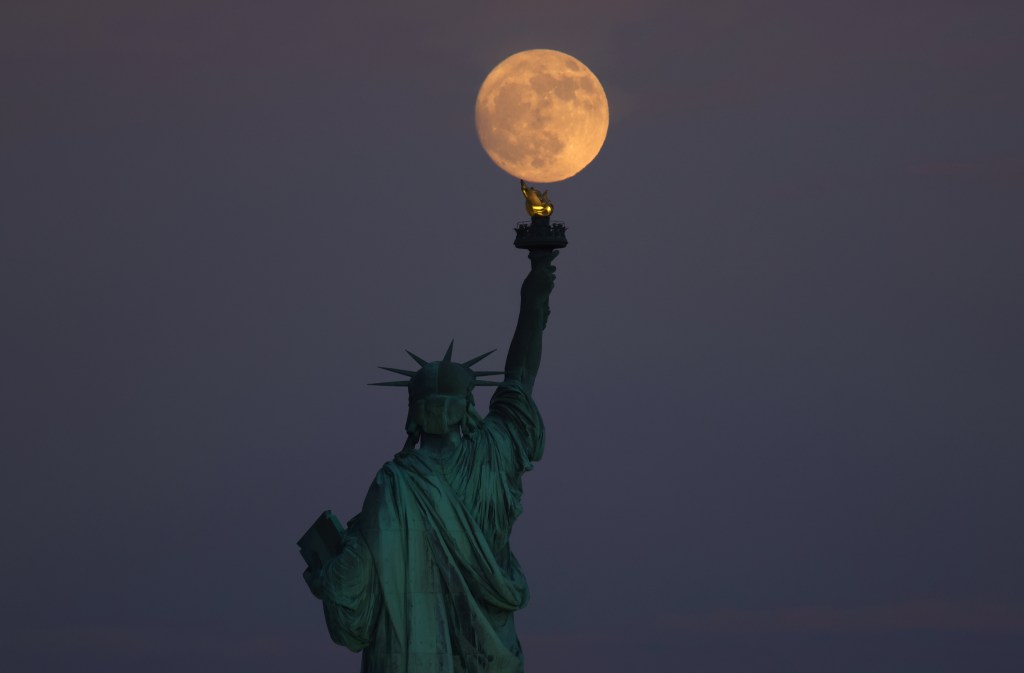 JERSEY CITY, NJ - JUNE 20: The Strawberry Moon rises behind the Statue of Liberty as the sun sets on the summer solstice in New York City on June 20, 2024, as seen from Jersey City, New Jersey.  