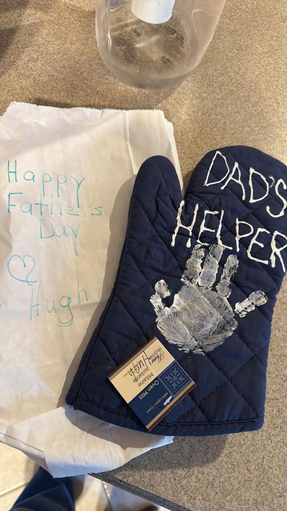 An oven mitt with the message "Dad's Helper" on it, along with a child's hand print painted on it, as well as a note that says "Happy Father's Day" from Nagel's son Hugh