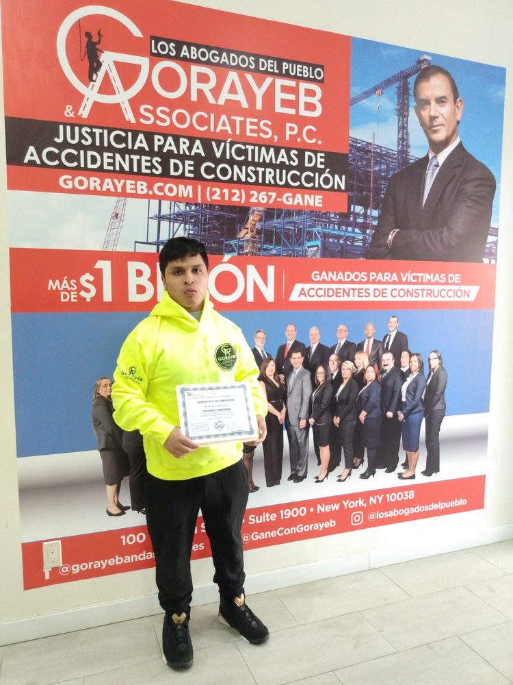 A man displays his FastTrack Training Safety Center certificate while wearing a Gorayeb & Associates sweatshirt and standing inform of signage provided by the law firm.

