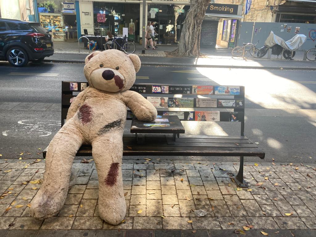 Larger stuffed teddy bear, bloody and battered, sits on a bench. 