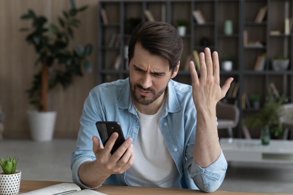 Angry young man holding a smartphone, frustrated by poor app performance and weak wifi signal