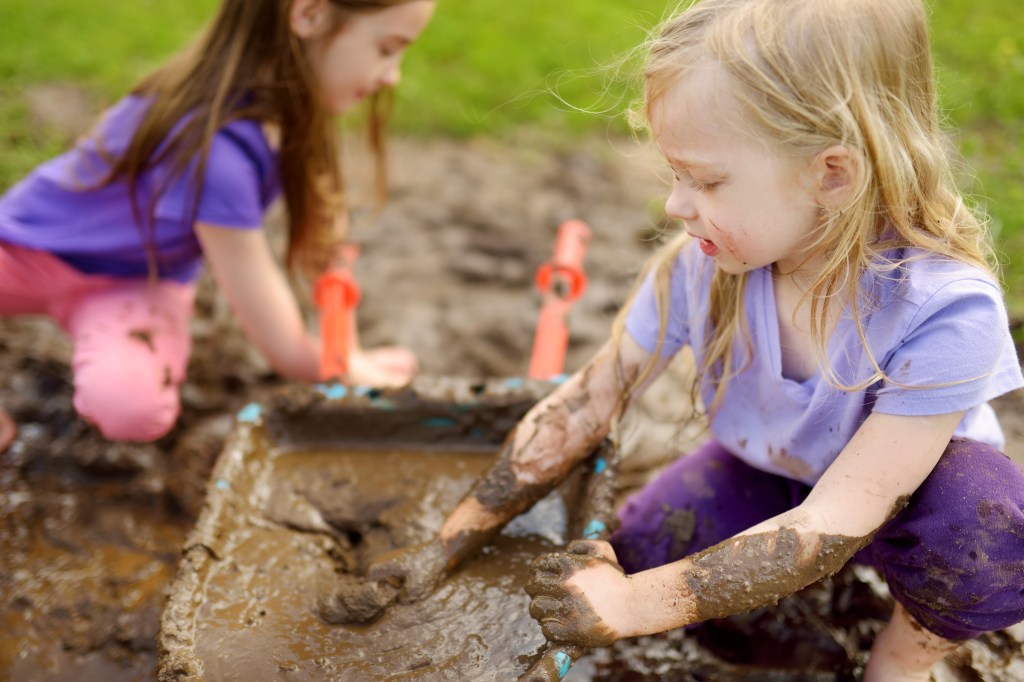 Two young girls joyfully playing and getting dirty in a large mud puddle outdoors on a sunny summer day