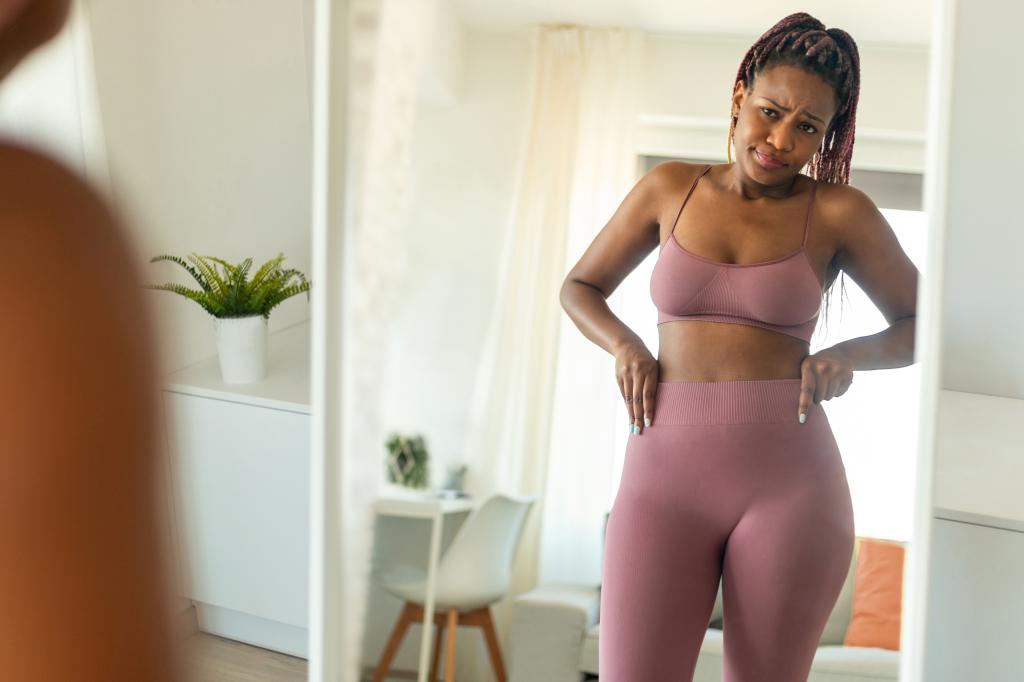 Upset black woman looking at her reflection in mirror, disappointed with her slimming diet results at home