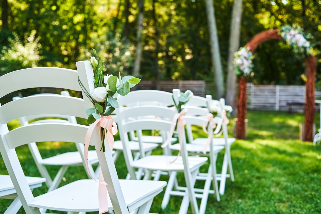 White wedding chairs adorned with fresh flowers and pink ribbons, placed outdoors on green grass for a wedding ceremony
