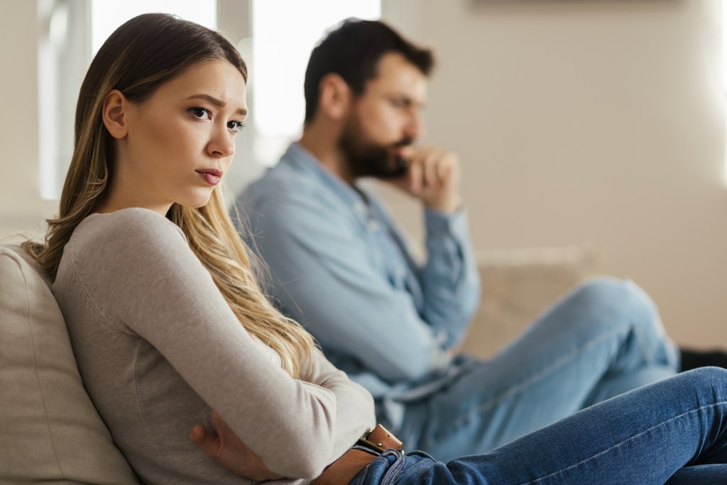 Worried young woman sitting on sofa at home with her partner ignoring her 