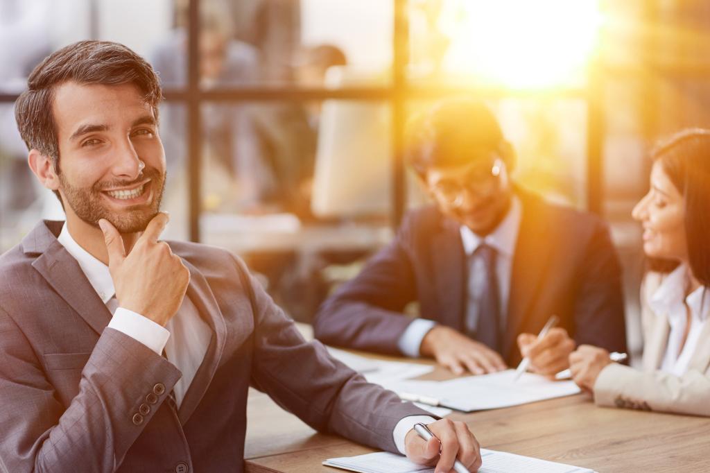 young man posing for the camera while sitting at a table in the office against the background of his colleagues