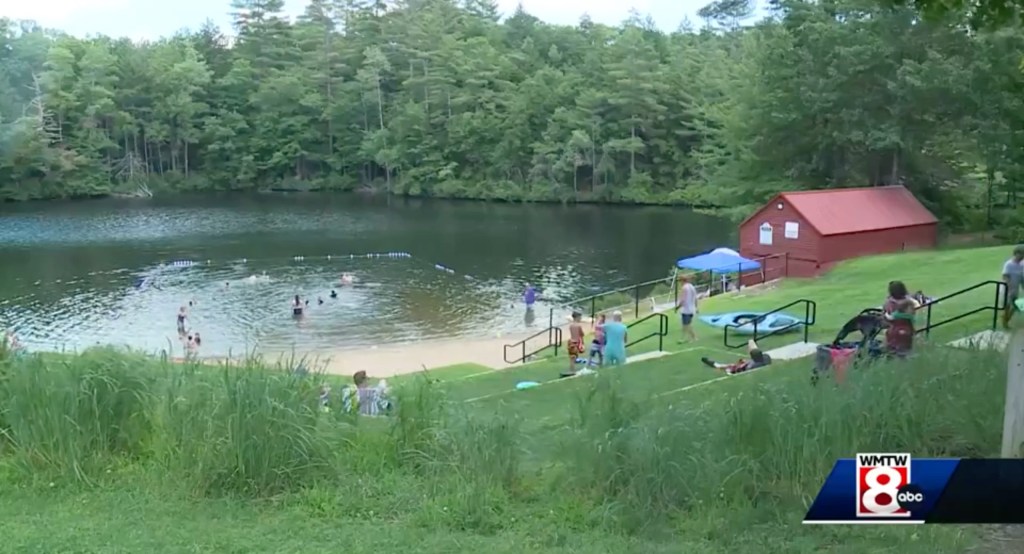 The Mousam River is surrounded by trees and grass with a few people swimming, and others sitting on a small hill