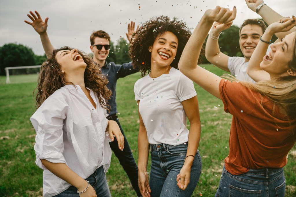 Group of five friends having fun at the park - Millennials dancing in a meadow among confetti thrown in the air - Day of freedom and carefree