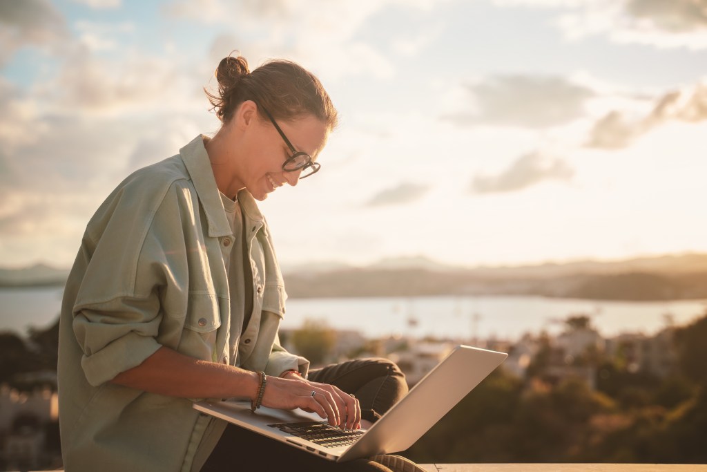 Beautiful young girl woman in eyeglasses sitting with a laptop on her balcony at sunset with a view of the city, remote work from home, a successful freelancer.