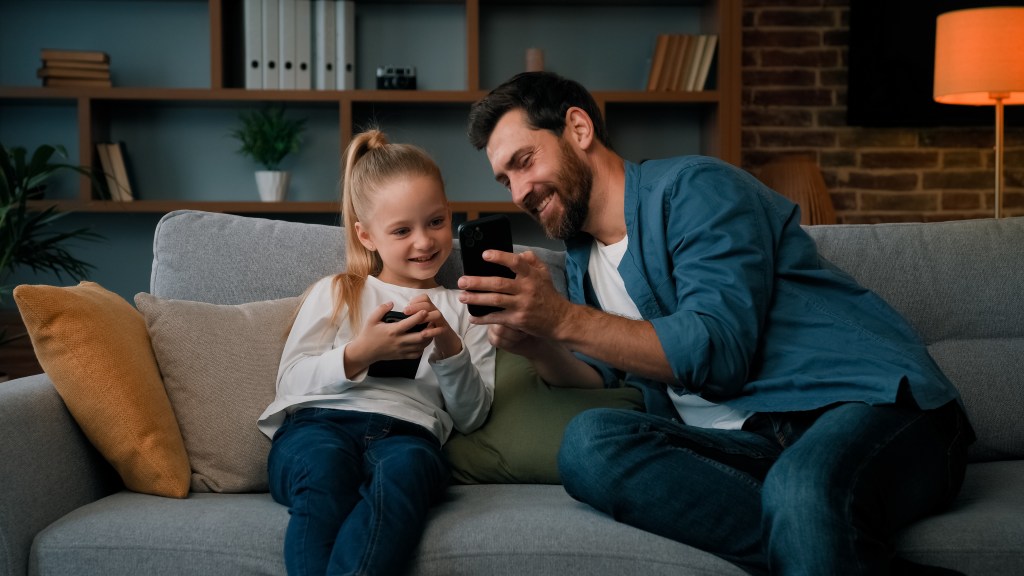 Caucasian man and young girl sitting at home, engaged with a smartphone, learning and exploring mobile apps together