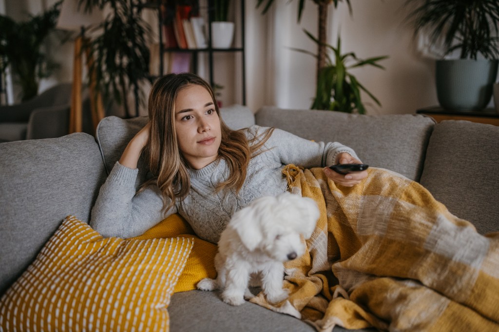 Young woman relaxing on a sofa while watching television with her Maltese puppy.