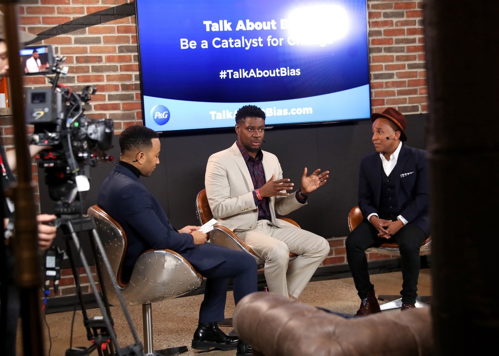 Three men sit on chairs in front of a screen that says "Talk About (blurred) Be a Catalyst for (blurred) #TalkAboutBias. A video camera tapes the men. 