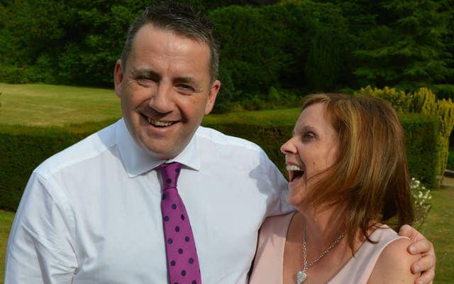 John Hunt, a BBC sports commentator, is seen wearing a white Oxford shirt and  pink tie, while smiling and wrapping his left arm around the shoulder of his wife, Carol, a woman with mid-length brown hair seen laughing in the photo.