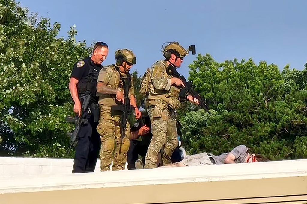 a uniformed cop and 3 men wearing army fatigues and holding rifles stand over the shooter's dead body.