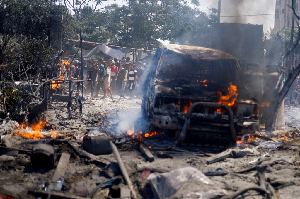 Palestinians gathered near damages at a tent camp in Al-Mawasi, Khan Younis, following an Israeli strike, with a car on fire in the background