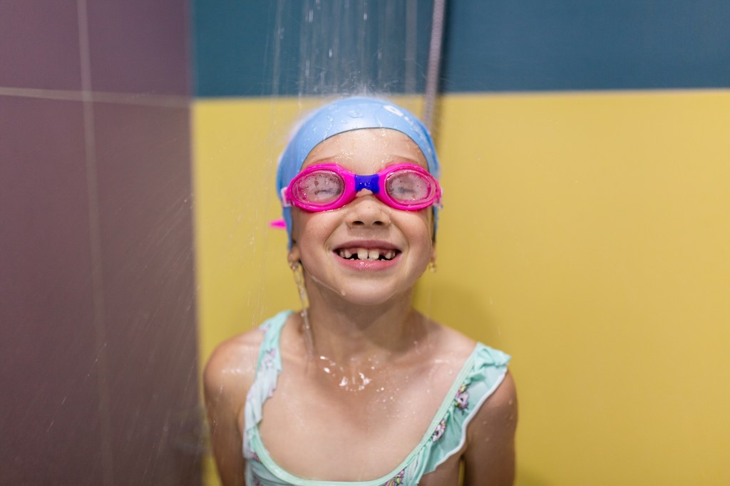 Smiling little girl swimmer with goggles and swimwear looking at camera while taking a shower after workout in pool