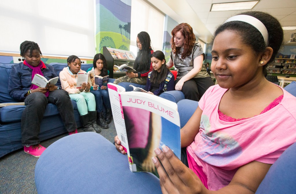 A girl reads a Judy Blume book in a library, other children on a couch behind her in the distance.