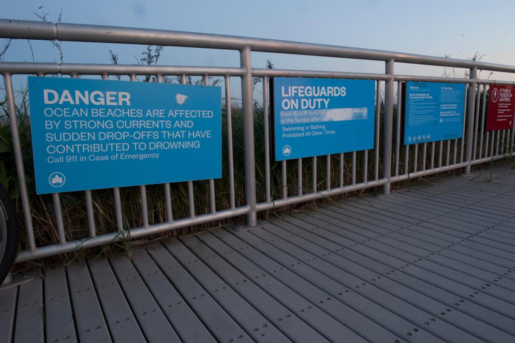 Danger signs on boardwalk in Far Rockaway saying strong currents contributed to drownings and lifeguards on duty.