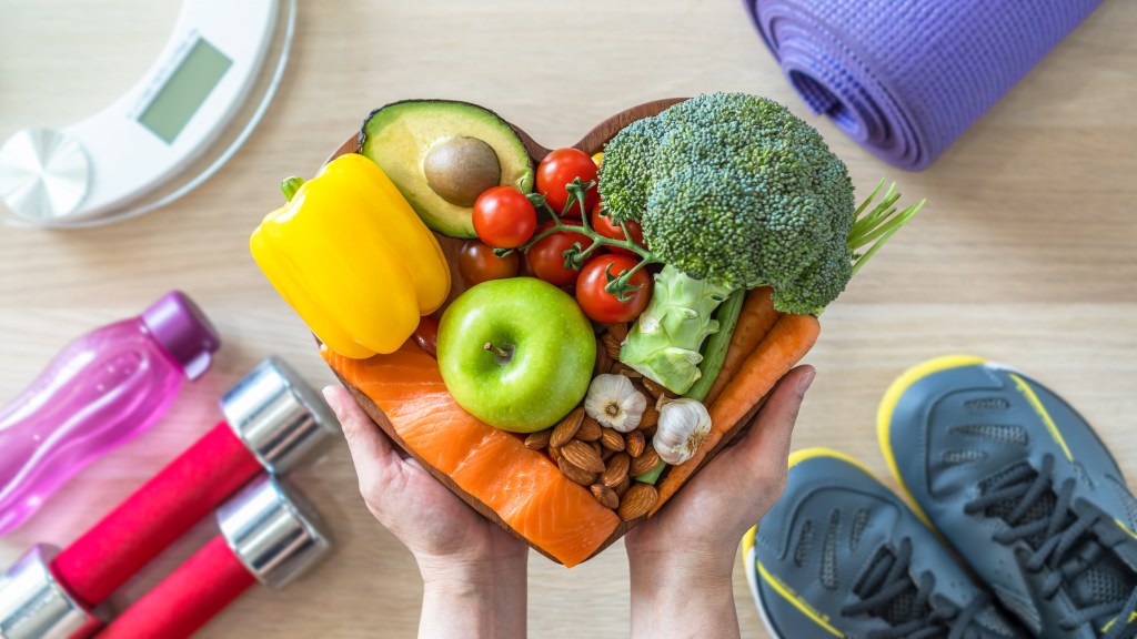 Heart-shaped bowl filled with healthy ketogenic diet food, gym workout equipment, sports shoes and weight scale in a fitness center