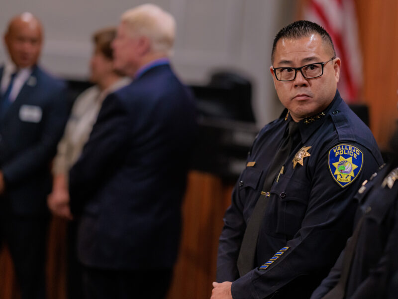 A focused police chief in uniform with the Vallejo Police badge visible, standing with his hands clasped in front of him. In the softly blurred background, several individuals are conversing, wearing professional attire. The setting is inside a city council chambers during a press conference at which California Attorney General Rob Bonta announced a lawsuit and consent decree against he Vallejo Police Department.