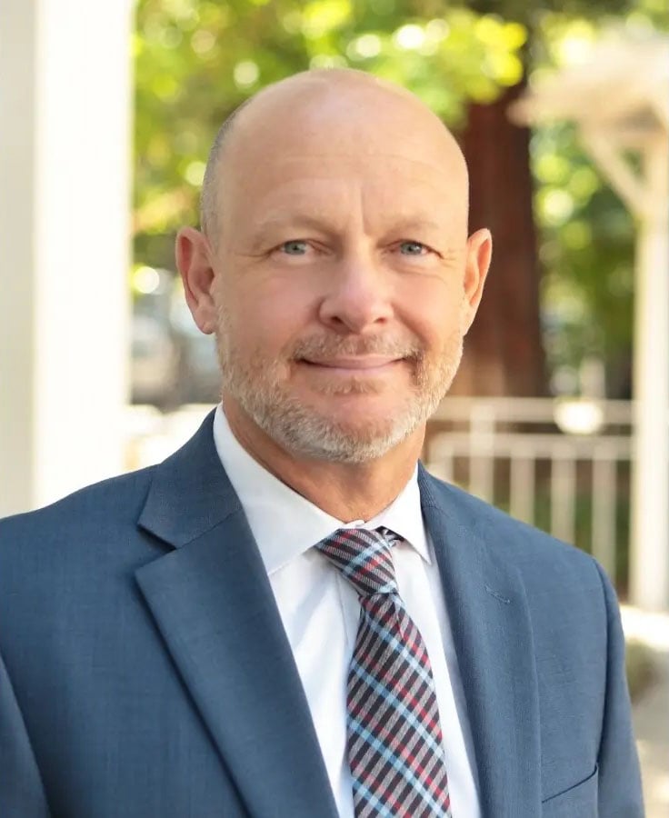 A headshot of a bald man with a close-cropped beard, smiling at the camera, wearing a suit, a light blue shirt, and a plaid tie. He appears professional and approachable, standing against a blurred natural background.