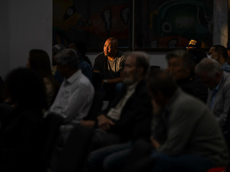 A man bathed in warm sunlight listens to a speaker at a community event while surrounded by a diverse group of community members.