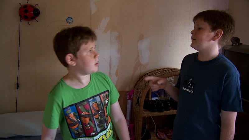 Two boys face each other, one pointing to the other with lips parted in mid-speech. Ladybug hangs on wall with peeling paint behind them.