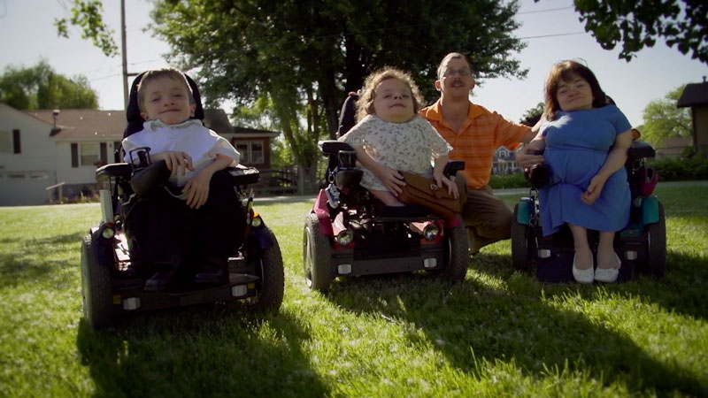 The Davert family looks at us and smiles in their yard. Three of the family members are using wheelchairs and have dwarfism from Brittle Bones Disease. The fourth family member, an older man, squats in between the rest of his family members.