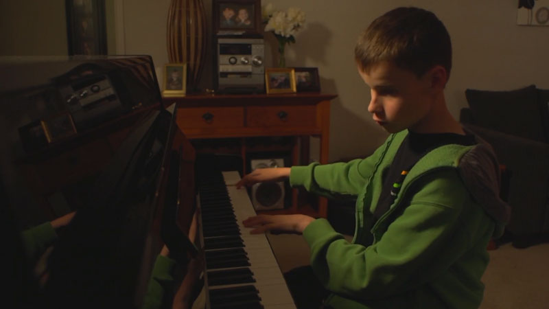 Cam, a young blind boy, wears a green jacket and sits in front of a grand piano in his house. He places his hands gracefully on the keys as he plays the piano.