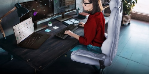 A woman working on a desk with a Lenovo ThikStation P1 Gen 7.  She also has 2 monitors, a keyboard, and a mouse.  At the back, a pile of books and a plant can be seen.