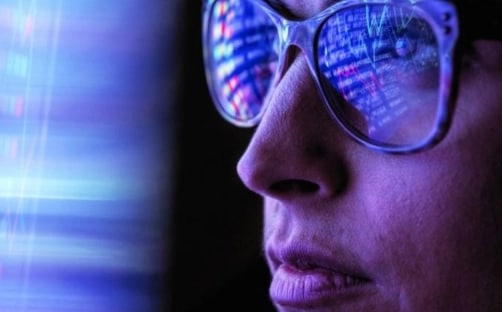 A closeup of a person wearing glasses, looking at digital screen, with the information on the screen reflecting in their glasses.