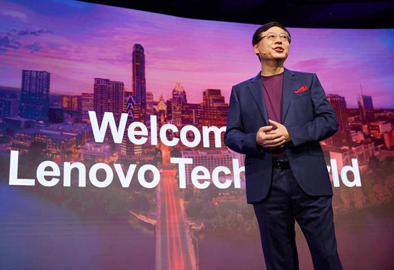 A man in a suit speaks at Lenovo Tech World with a cityscape background and “Welcome” text displayed.