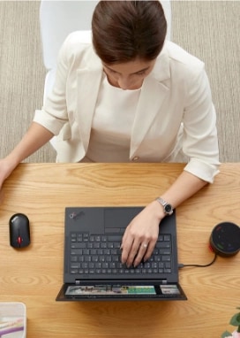 Shot from above, a woman sits at her desk while drinking coffee and shopping online with her Lenovo ThinkPad laptop.