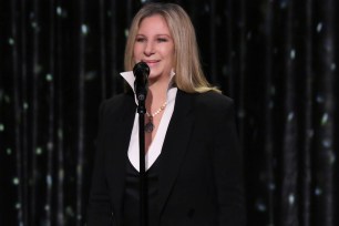 Barbra Streisand, dressed in a black suit, standing at a podium.