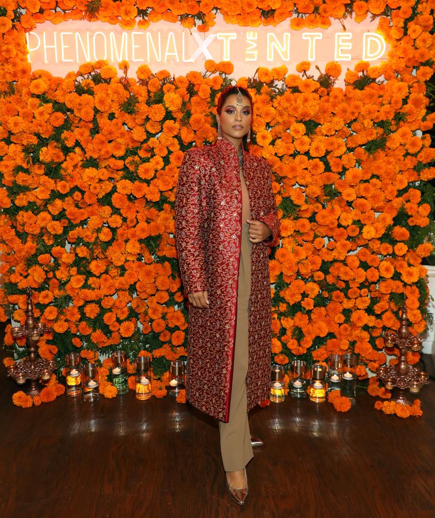 Lilly Singh posing in front of a wall of orange flowers.