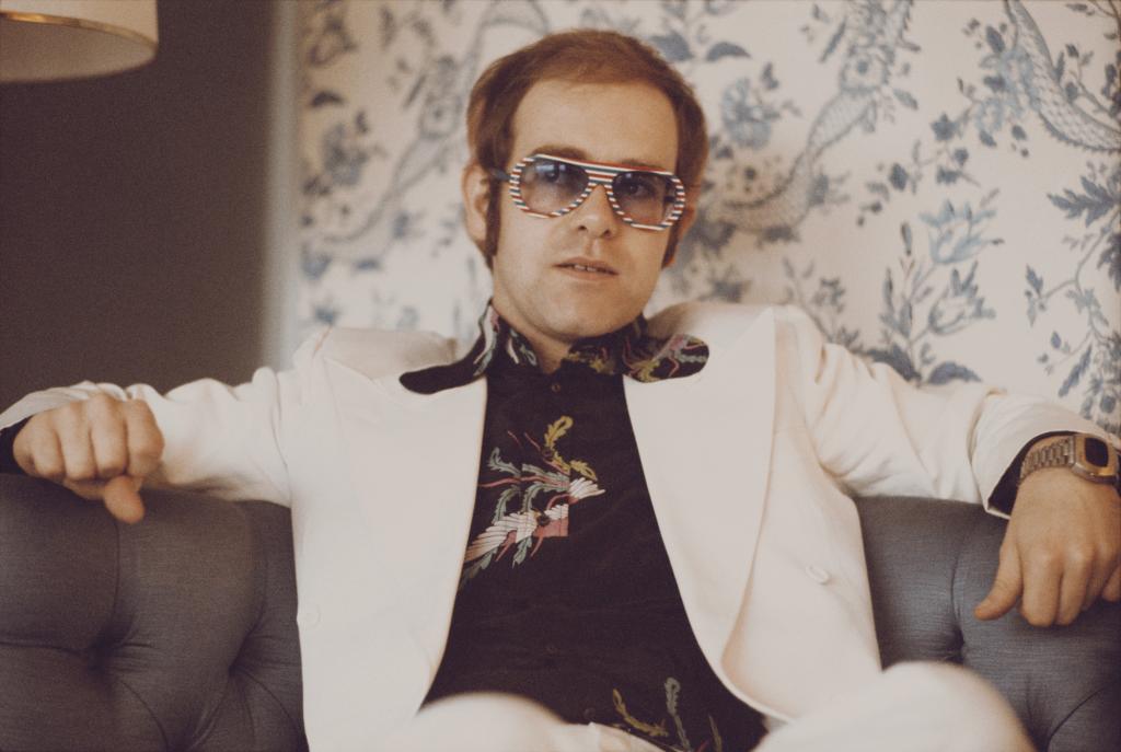 Elton John, wearing a white suit, black shirt with flower motif and multicolored sunglasses in 1973.