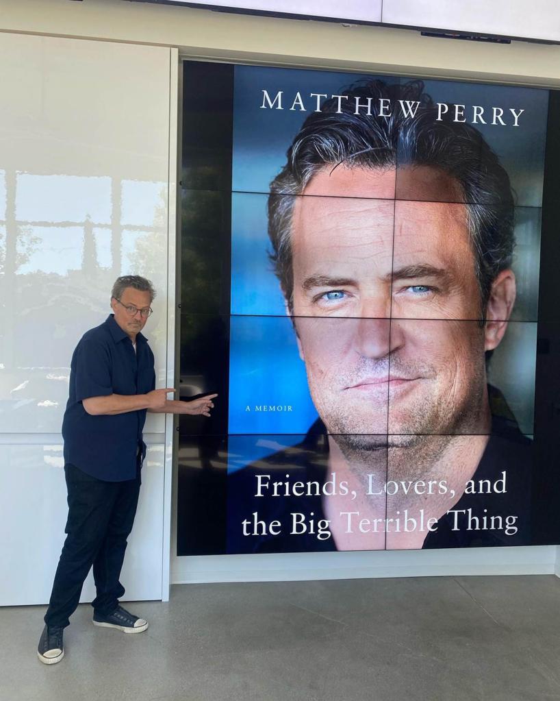 Matthew Perry posing next to a big photo of the cover of his memoir.